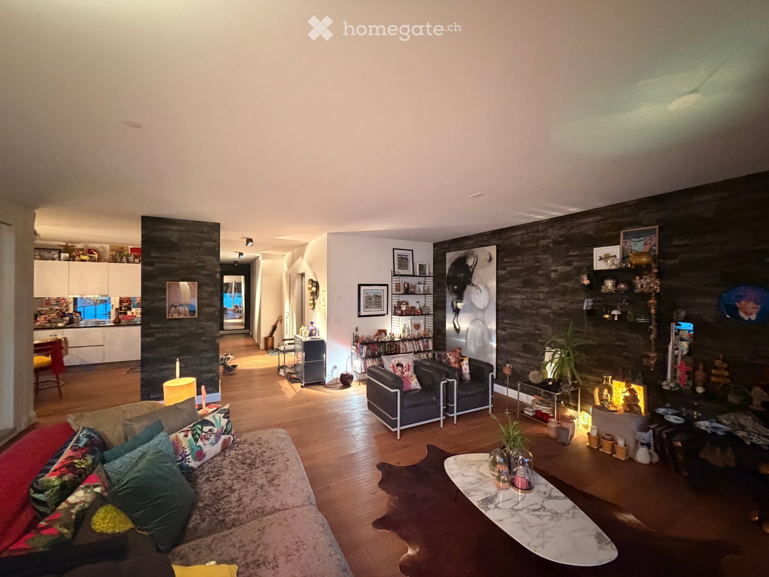Living room with wooden flooring, a large sofa with colorful cushions, a coffee table, a side table, shelves with decorative items, and framed pictures on the wall.