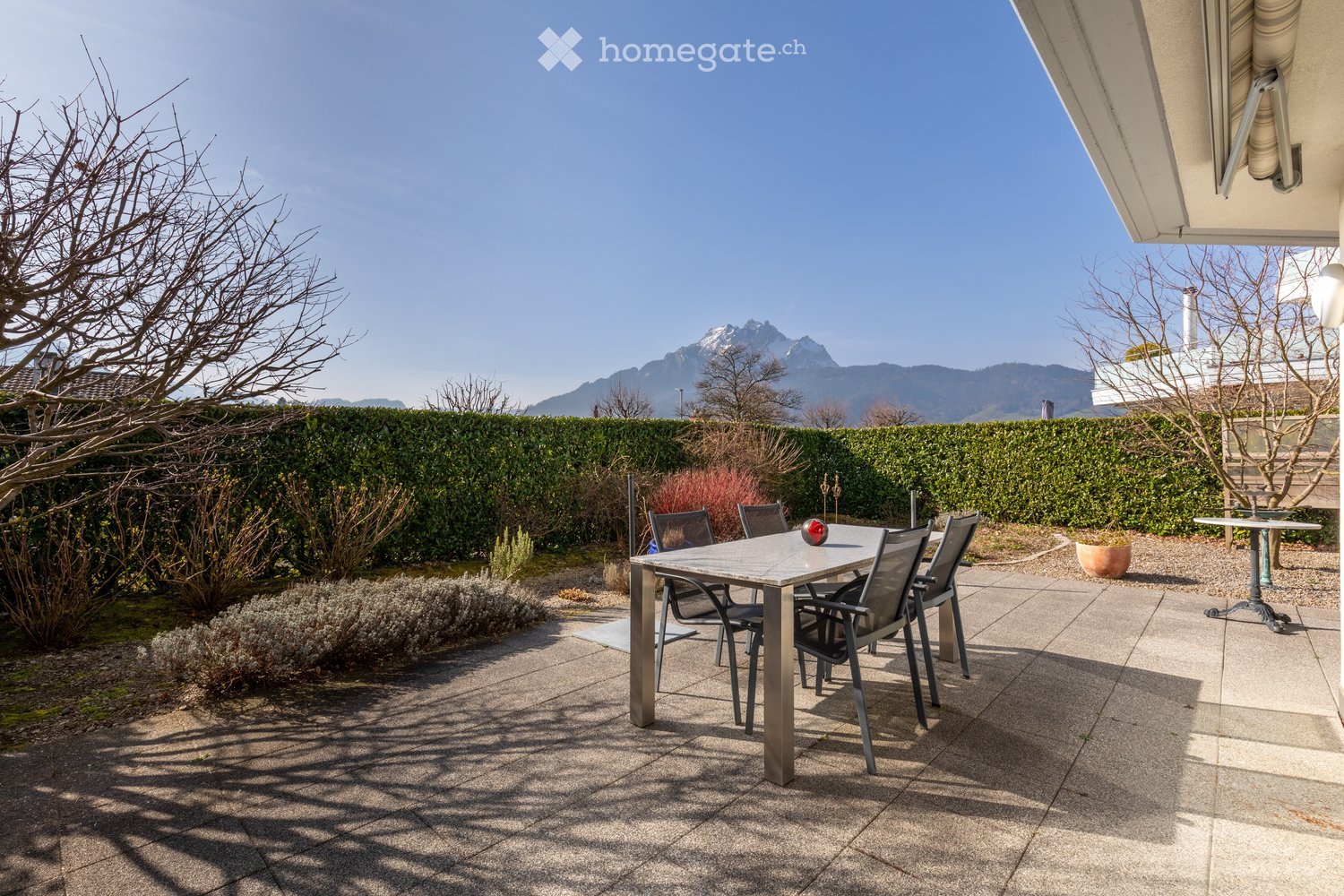 Outdoor patio with a table and chairs, surrounded by a hedge and a view of mountains.