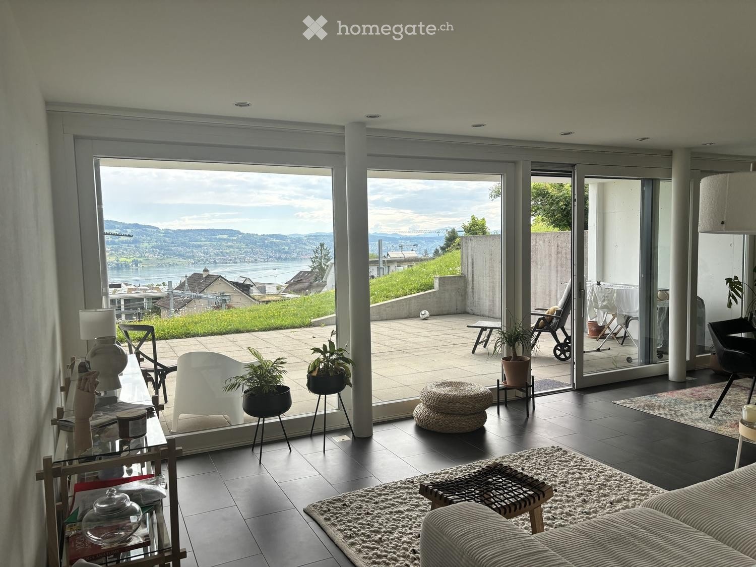 Modern living room with large glass doors opening to a balcony, offering a scenic view of a lake and hills. The room features a sofa, coffee table, chairs, a lamp, and potted plants.