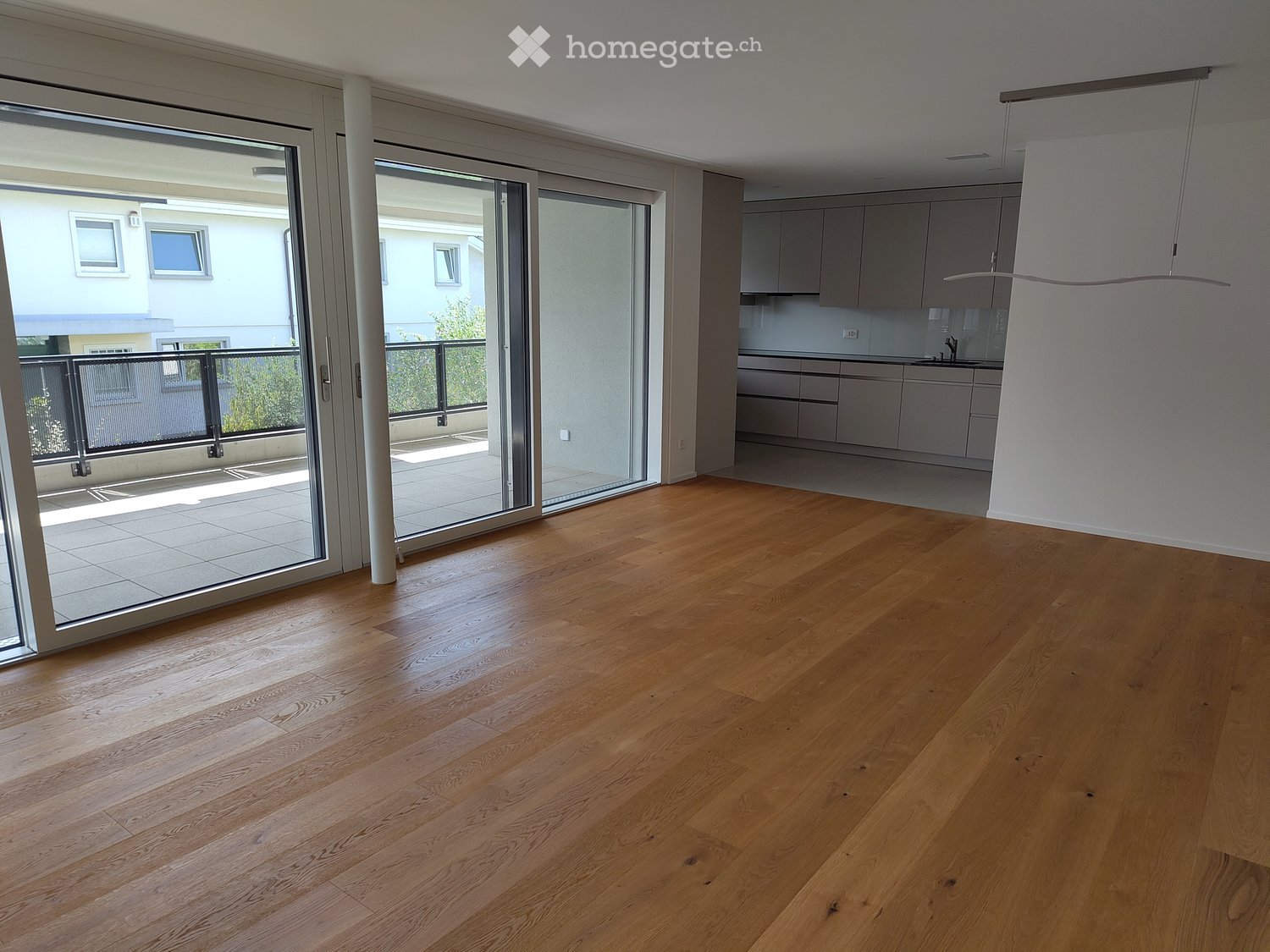 Empty room with wooden flooring, large sliding glass doors leading to a balcony, and a modern kitchen area visible in the background.