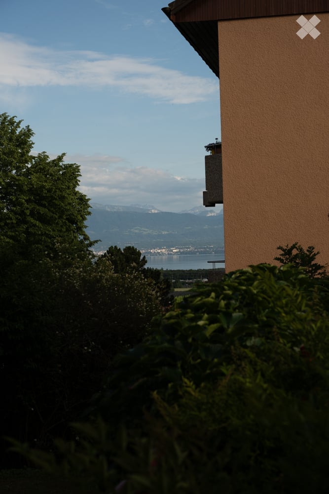 Balcony with a scenic view of mountains, a lake, and a town in the distance, surrounded by greenery.