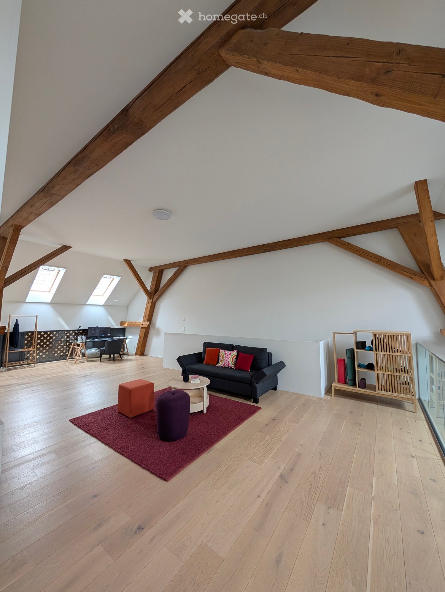 Bright, open living space with wooden beams, hardwood floors, and large windows. Features a black sofa with colorful cushions, a coffee table, two stools, a shelf, and a small workspace area with a laptop.