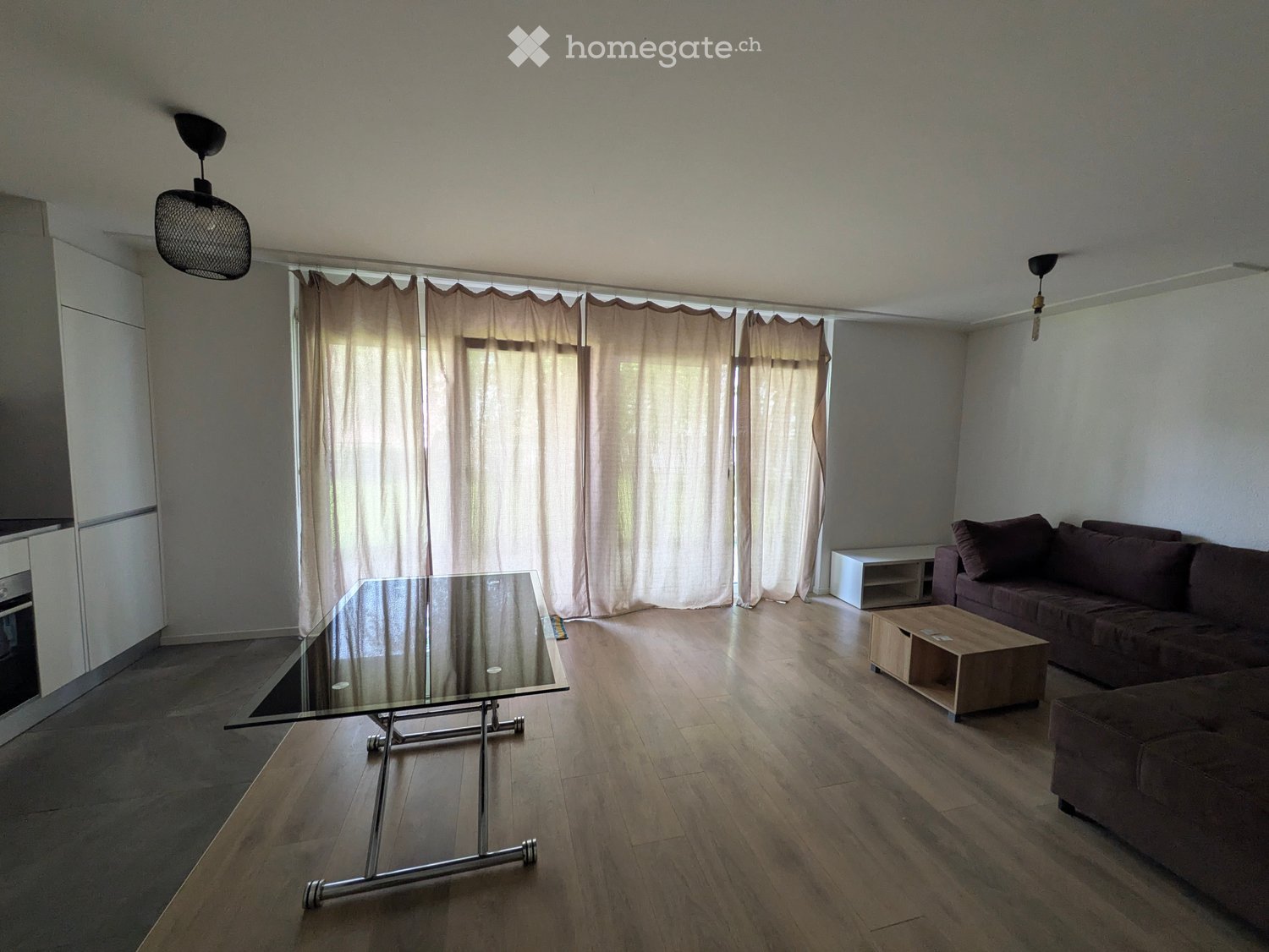 Modern living room with wooden flooring, large windows covered by beige curtains, a glass coffee table, a dark brown sofa, and a small wooden side table.