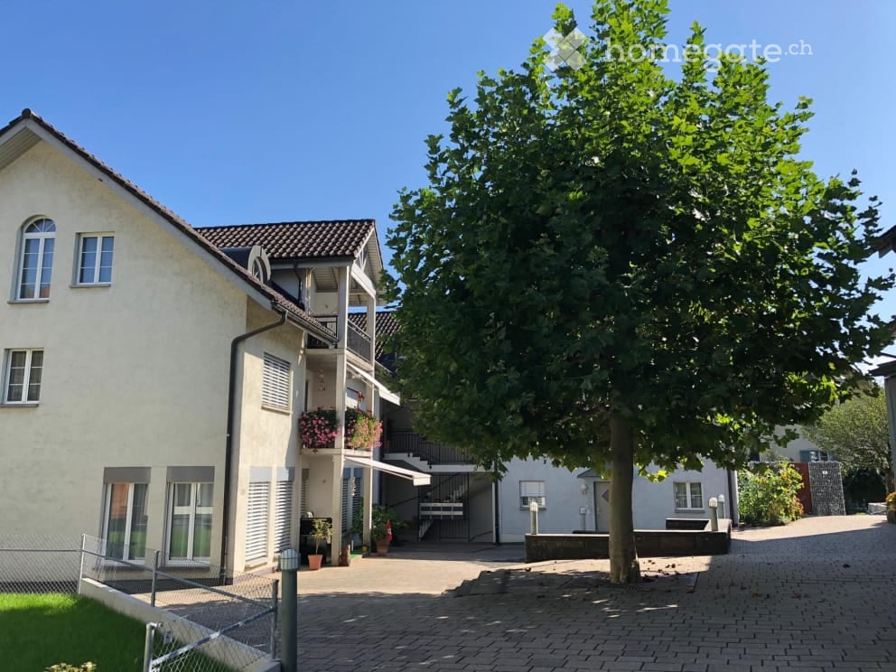 Two-story residential building with white exterior, multiple windows, and a balcony with flower boxes. The building is surrounded by a paved area with a large tree and some potted plants.