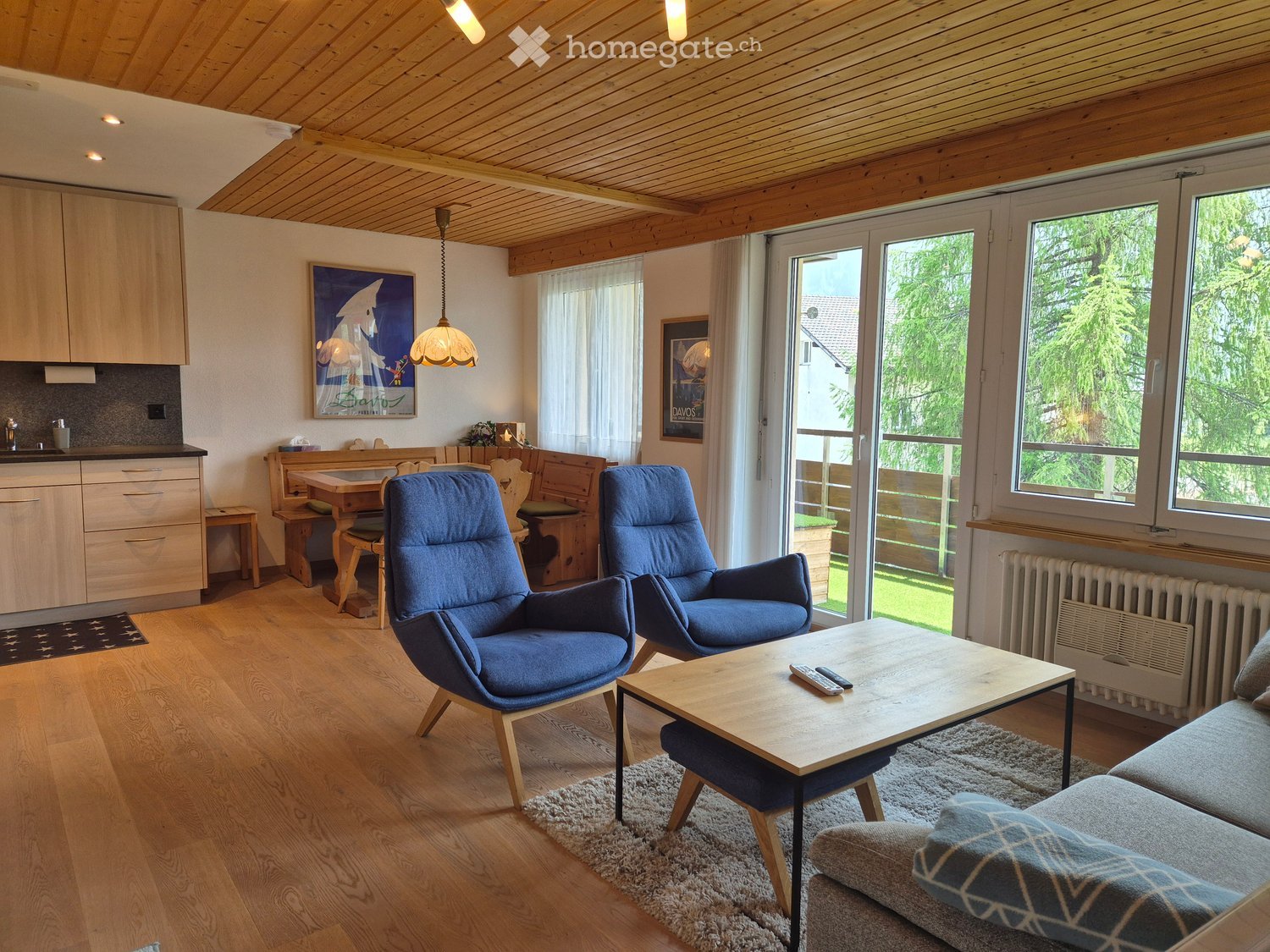 Living room with wooden floors, a sofa, two armchairs, a coffee table, a dining area with a table and chairs, and a kitchen area with cabinets and a countertop.