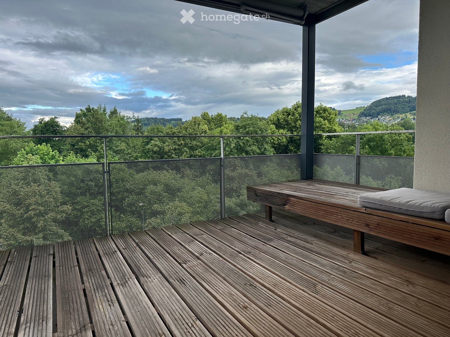Wooden deck with a bench and cushion, overlooking a green landscape with trees and hills under a partly cloudy sky.