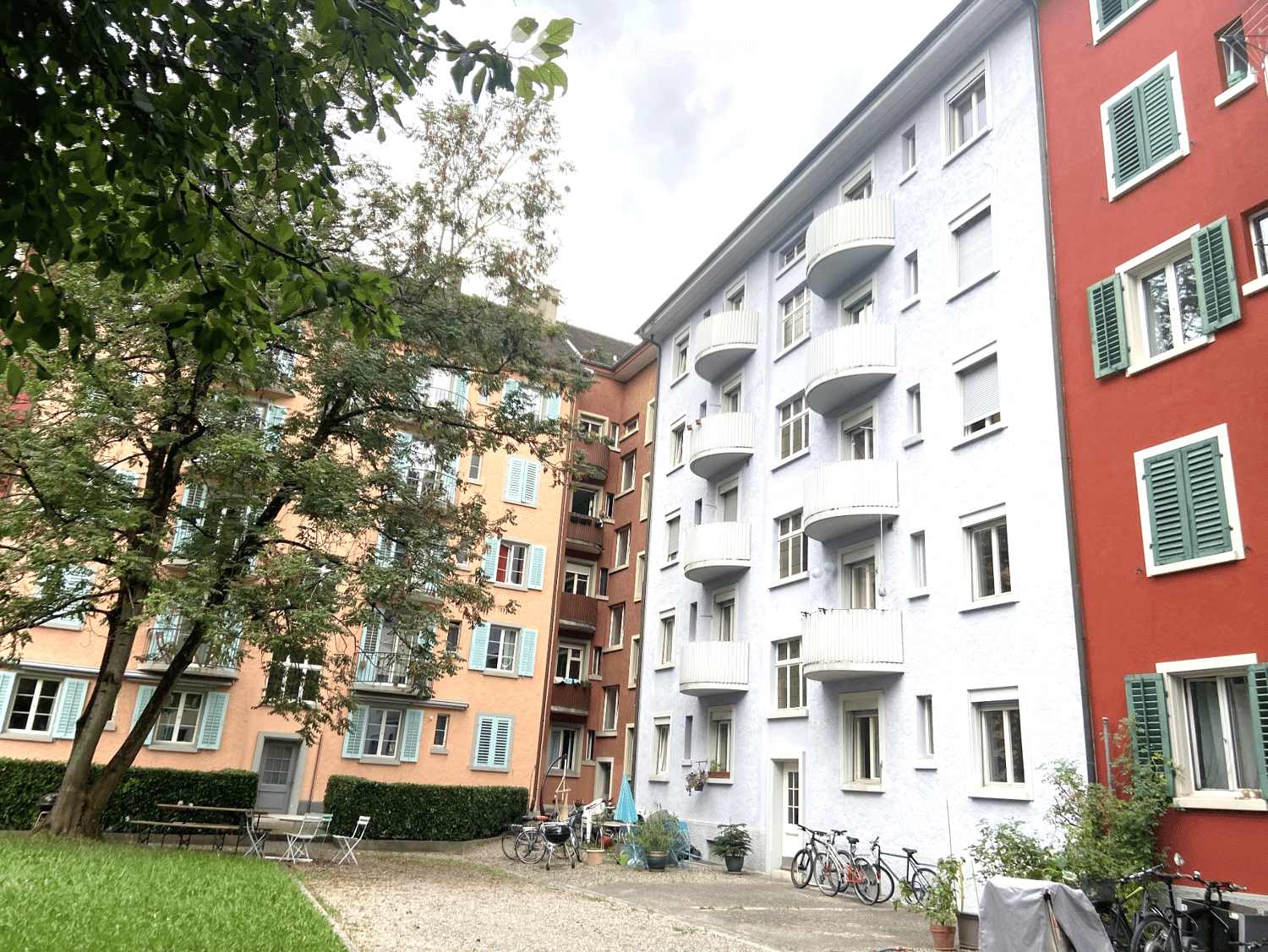 Three multi-story buildings with balconies, bicycles parked in front, greenery and trees in the area