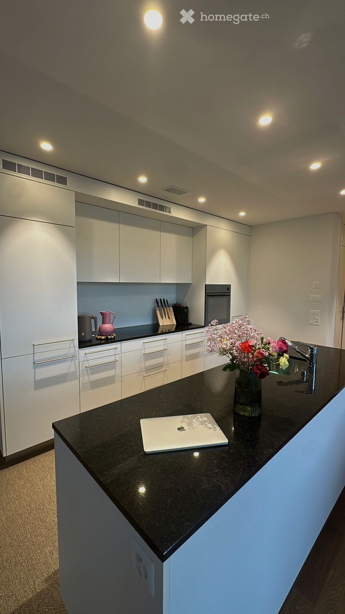 Modern kitchen with white cabinets, black granite countertop, laptop, flower vase, coffee maker, knife block, and oven.