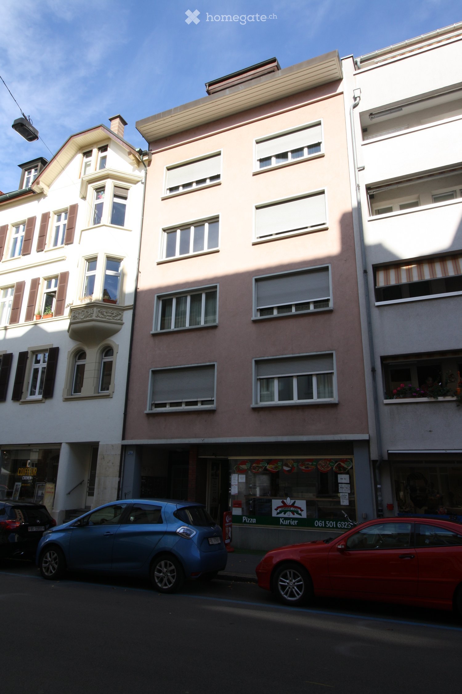 Three-story residential building with multiple windows, shutters, and a small balcony on the left side. A storefront with signage for a pizza and kurier service is visible on the ground floor, with several cars parked in front.