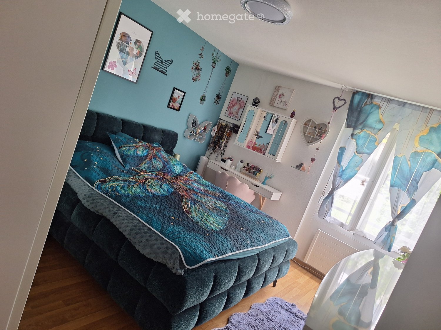 A cozy bedroom with a large bed, blue bedding, a nightstand, a mirror, and decorative picture frames on the walls. The room features wooden flooring and large windows with blue curtains.