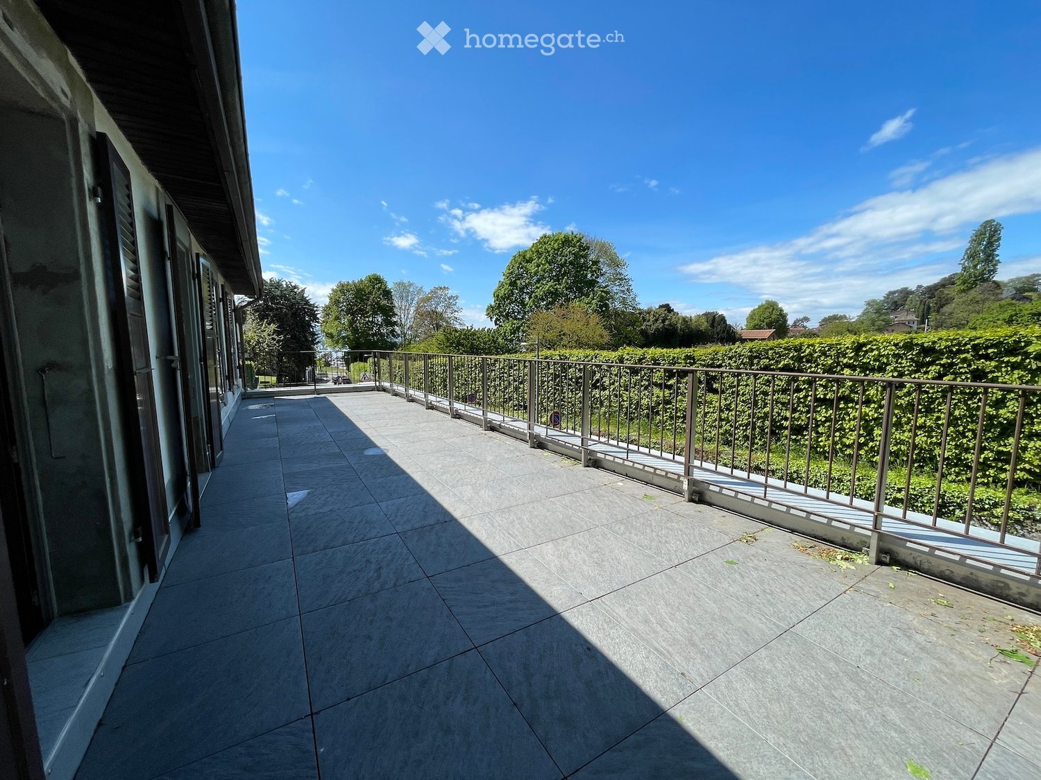 Balcony with tiled floor, metal railing, and greenery in the background under a clear blue sky.