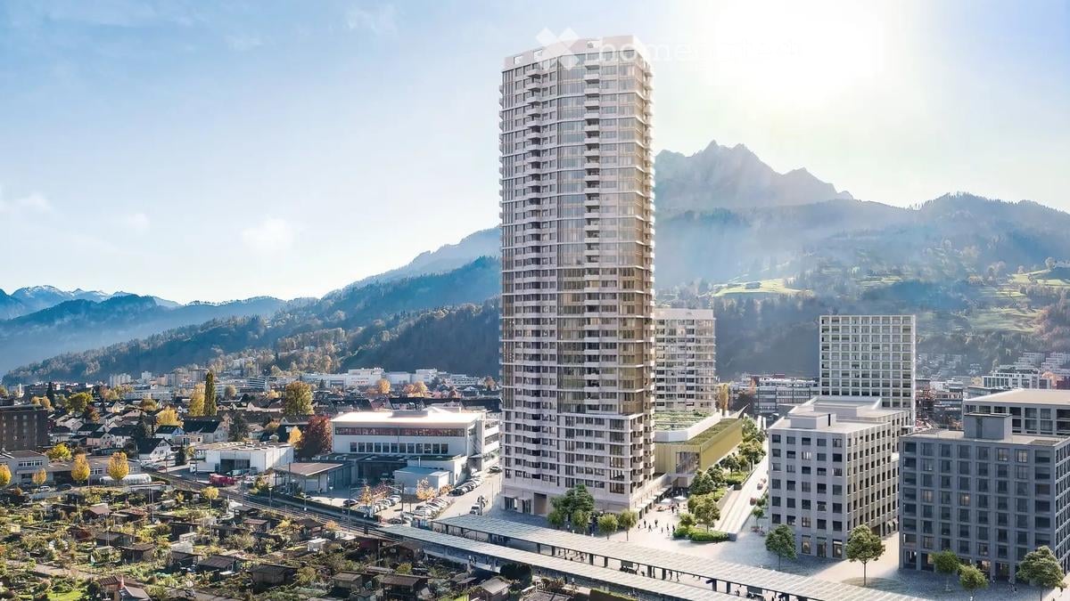Aerial view of a modern high-rise building surrounded by smaller structures and a scenic mountainous backdrop.