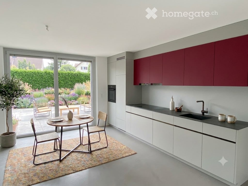 Modern kitchen with white and red cabinets, a black countertop, and a dining area with a round table and chairs. Large windows offer a view of a garden.