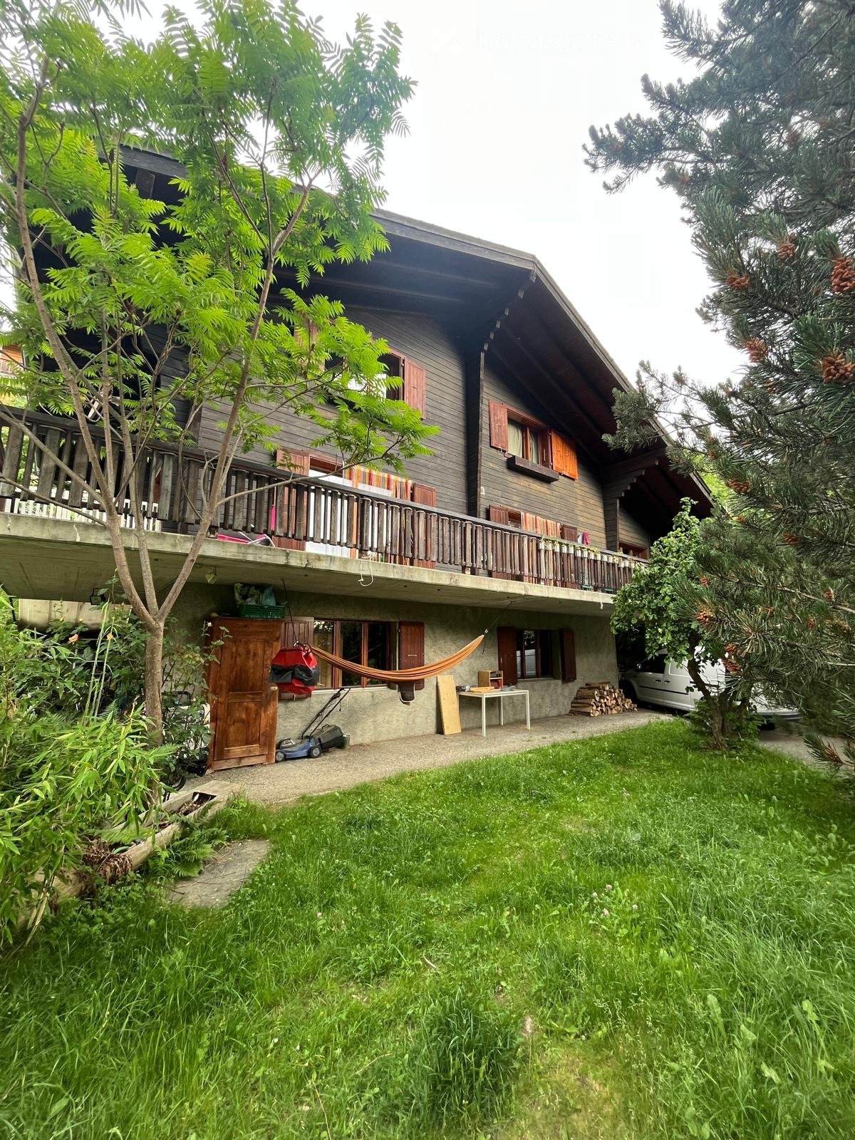 Two-story wooden house with a balcony, surrounded by trees and a grassy lawn. Features include a hammock, a small table, and a parked car.