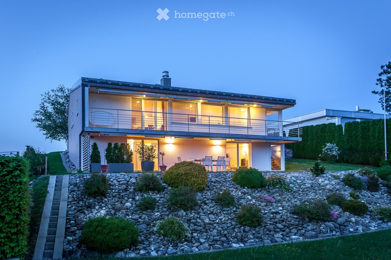 Modern two-story house with large windows and a raised ground floor, featuring a balcony and outdoor seating area.