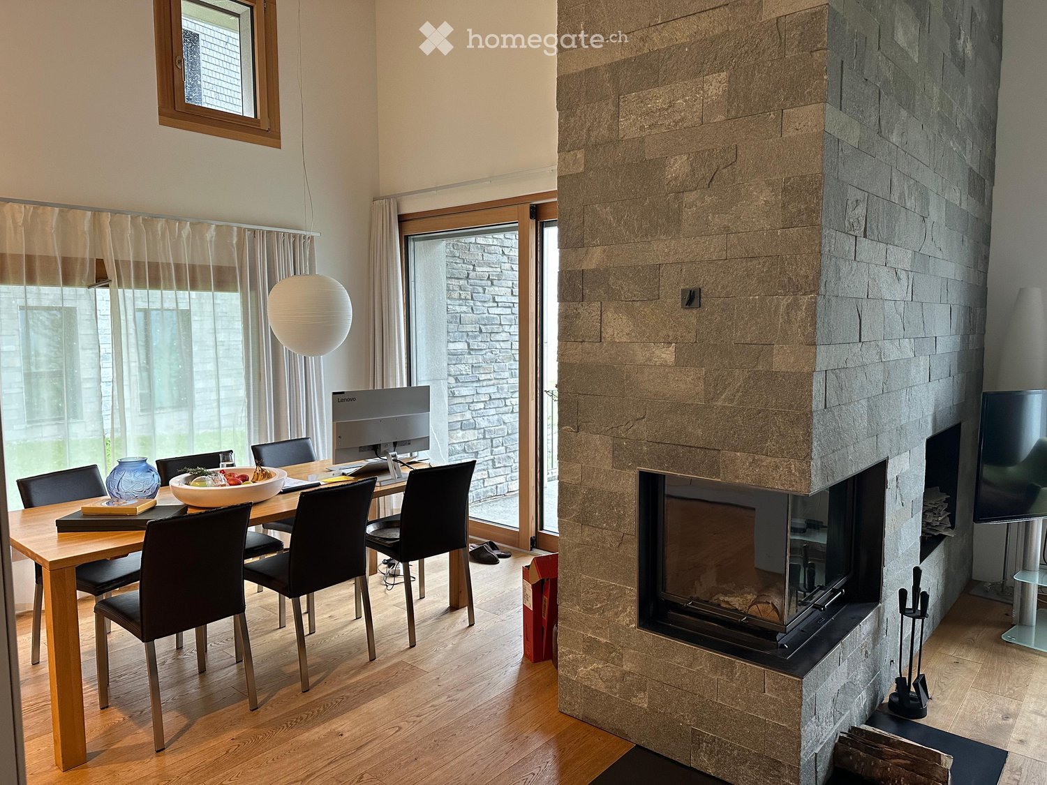 Modern living and dining area with a stone fireplace, wooden dining table, chairs, and a large screen. Natural light from large windows and a hanging ball lamp illuminate the space.