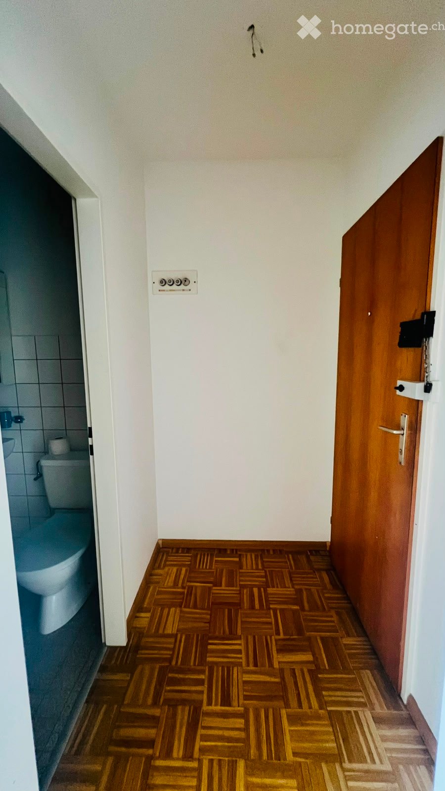 A narrow hallway with wooden parquet flooring and white walls, featuring a closed wooden door on the right and an open door leading to a bathroom on the left.