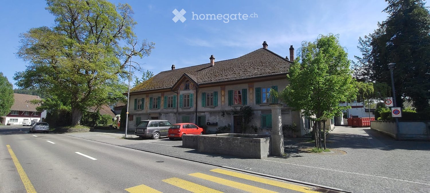 A two-story building with a pitched roof, green shutters, and a stone facade, situated on a street with parked cars and trees.