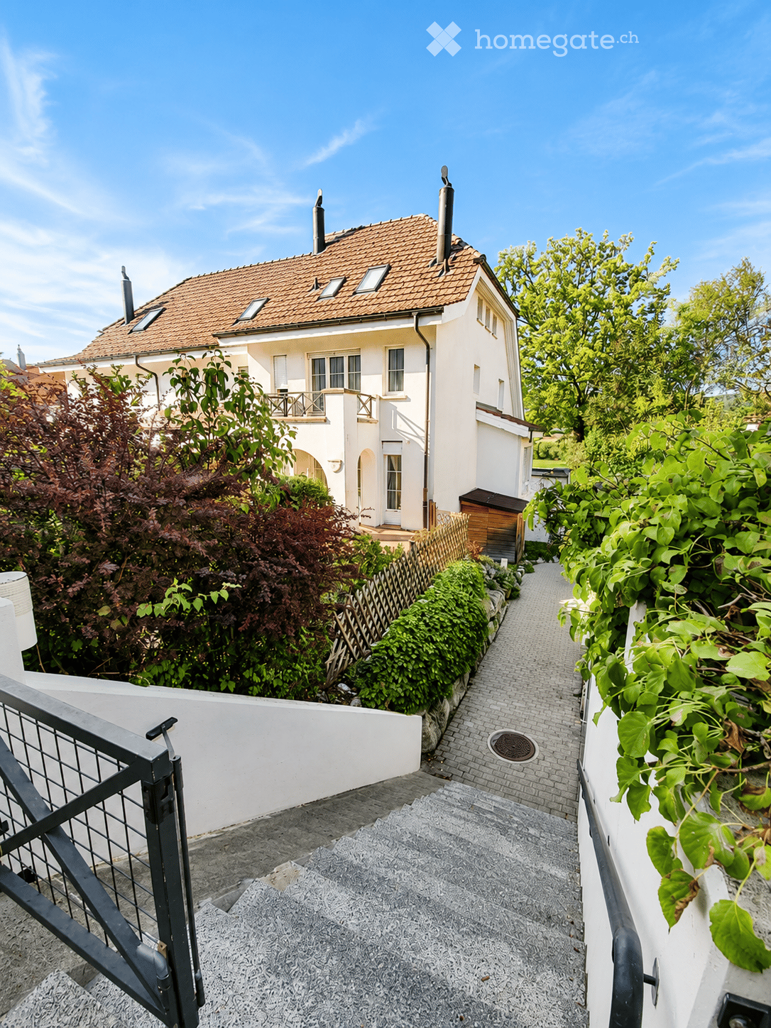 A two-story white house with a red-tiled roof, surrounded by greenery and a paved pathway leading to the entrance.