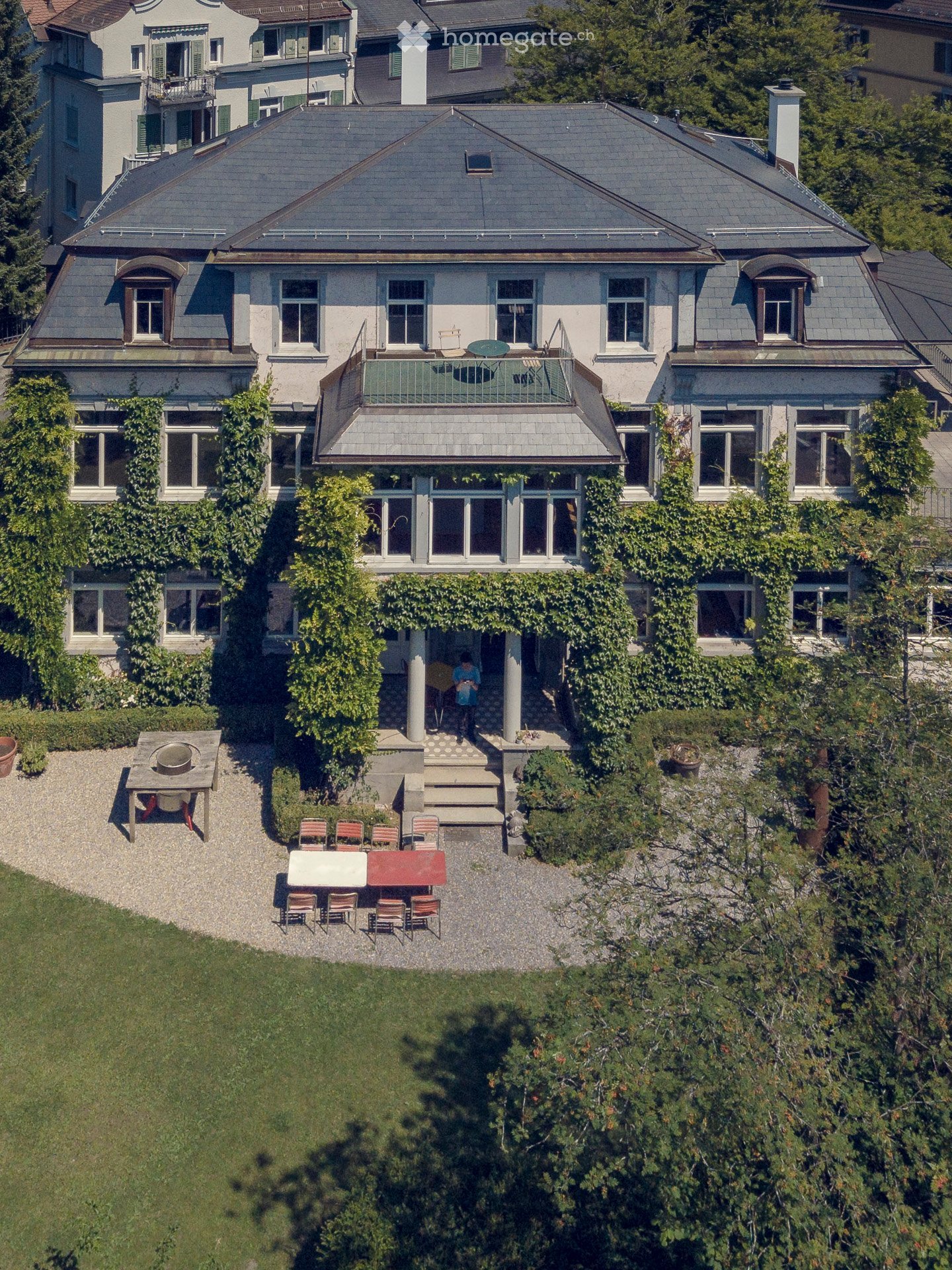 A multi-story house with a grey roof, white walls, and multiple windows. The house features a balcony with outdoor furniture and a well-maintained garden area with a patio and seating arrangement.