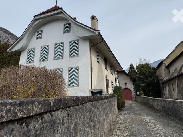 Traditional Swiss-style building with white facade, decorative black and white chevron-patterned windows, red door, and chimney. Surrounded by low stone wall and greenery.
