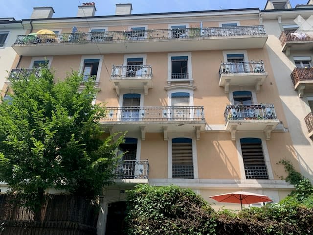 Multi-story residential building with multiple balconies and railings, light-colored facade, and greenery in the foreground.