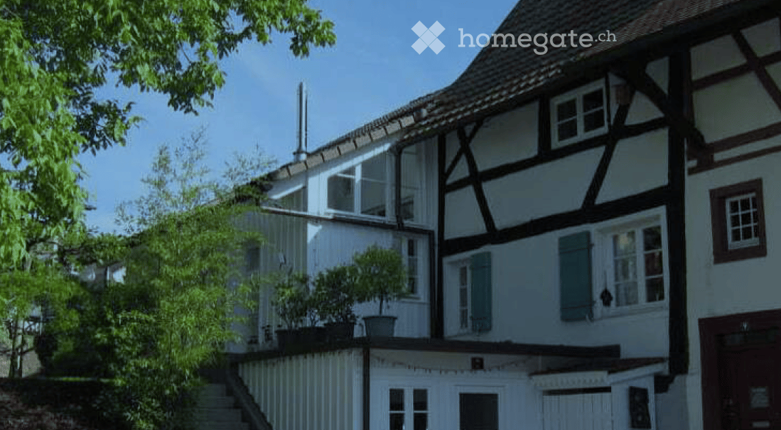 Traditional half-timbered house with white walls, dark wooden beams, multiple windows, and a sloped roof. A small garden with potted plants is visible in front.