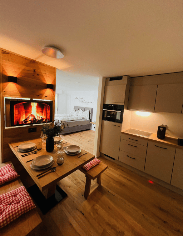 A modern, open-plan living and dining area with a central wooden dining table, fireplace, sofa, and a small kitchen area with cabinets and an oven.