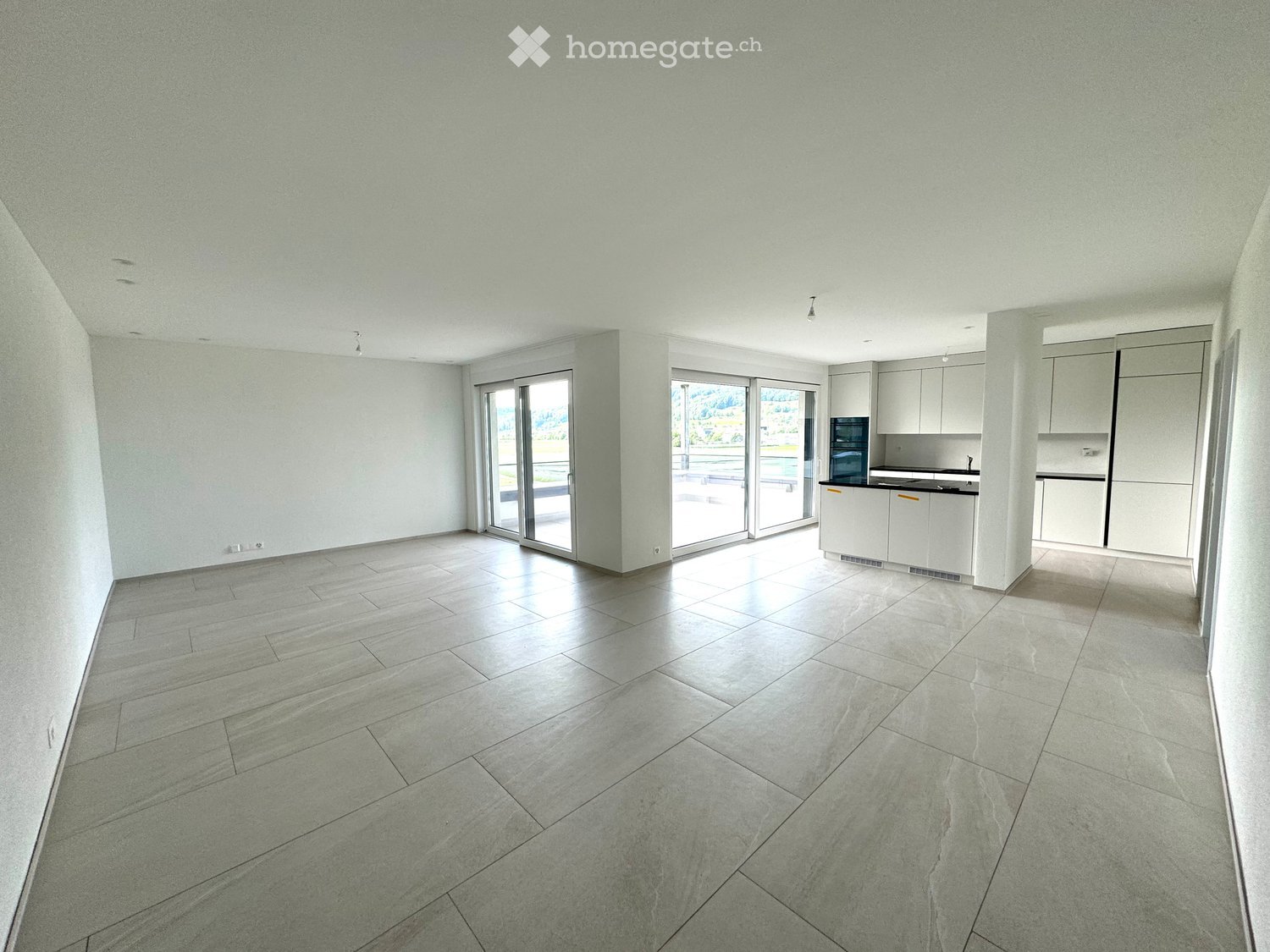 Empty room with light grey tiled floor, white walls, and large sliding glass doors leading to an outdoor area. Kitchen area with white cabinets and appliances is visible on the right side.