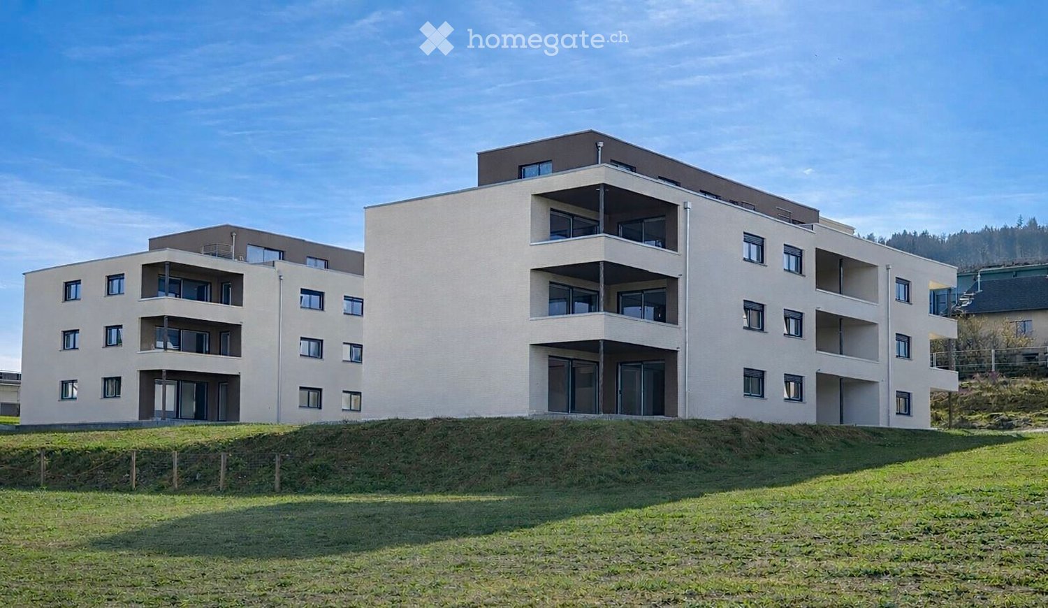Modern multi-story residential building with beige facade, multiple balconies, and large windows, situated on a raised ground floor with a grassy slope in front.