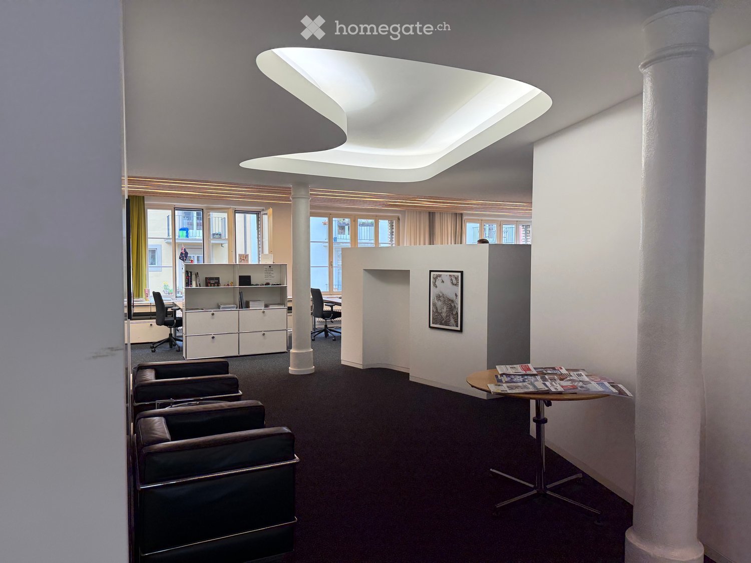 Modern office space with a reception area, featuring a sofa, chairs, a round table with magazines, a cabinet, and a picture frame on the wall.
