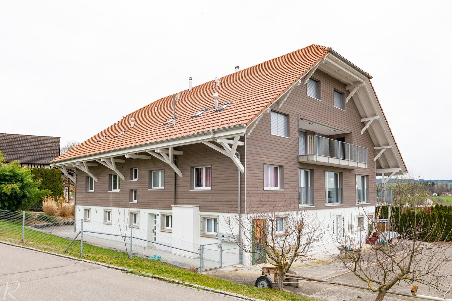 Two-story house with brown siding, red-tiled roof, multiple windows, and balconies on the upper level. The house is surrounded by a fence and some greenery.