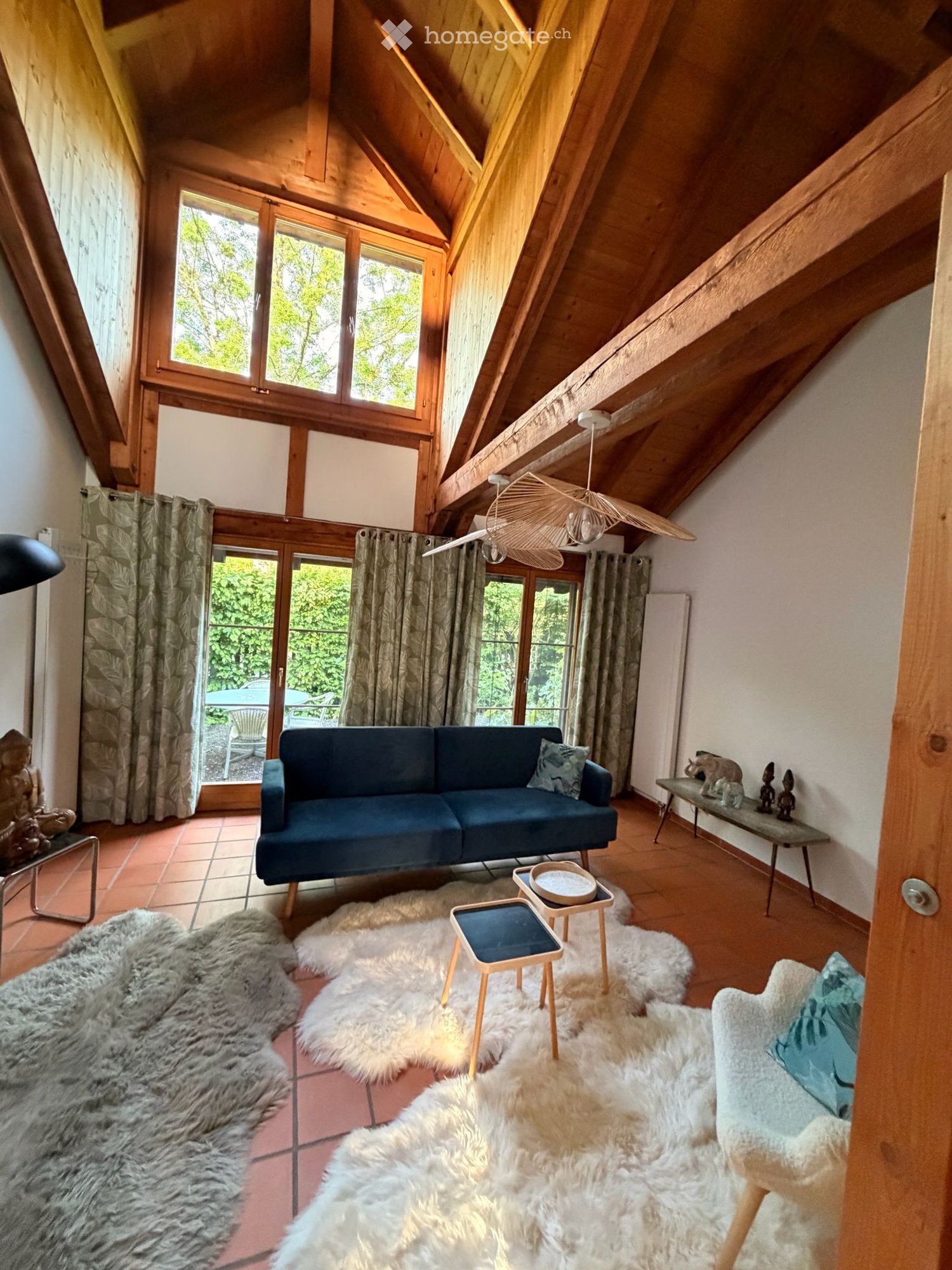 Living room with wooden beams, large windows, blue sofa, wooden stools, fur rugs, and decorative items on a side table.
