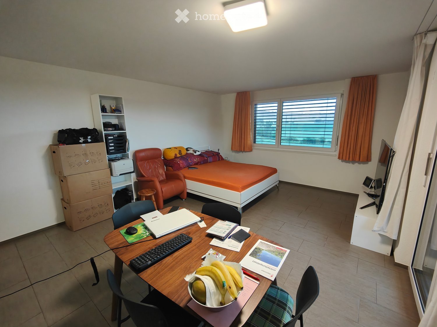 The room is furnished with a bed, a chair, a table with a laptop, keyboard, and mouse, and a screen. There are also curtains, a stool, and a shelf with items on it.