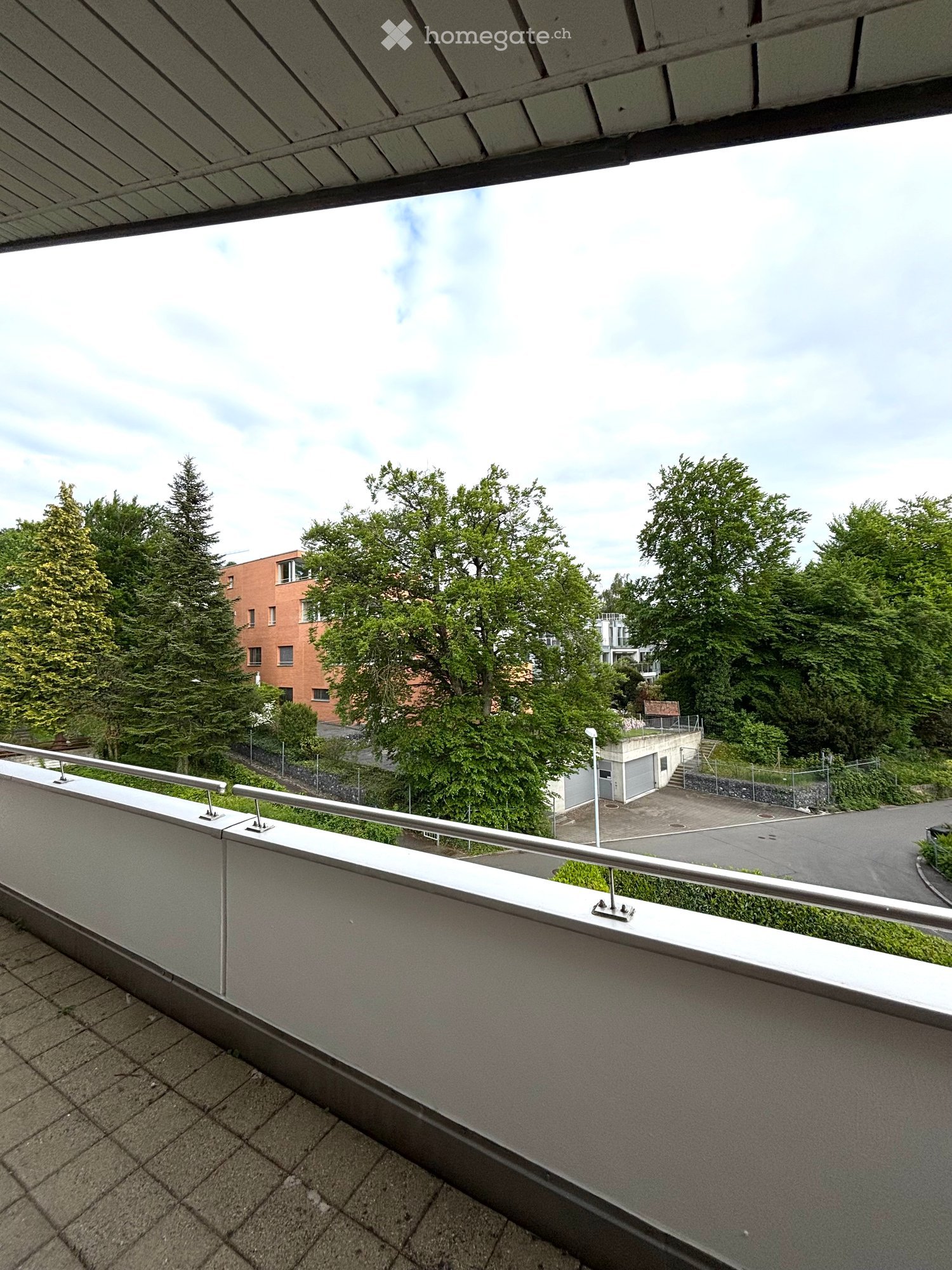 Balcony with metal railing, tiled floor, overlooking a parking area and greenery.