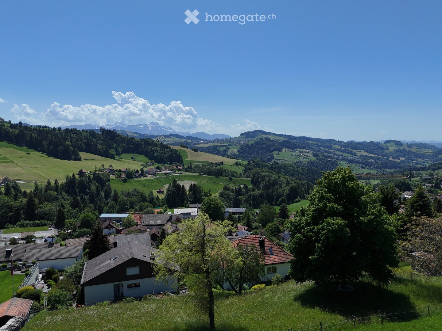 Aerial view of a rural village with scattered houses, lush green fields, and distant mountains under a clear blue sky.