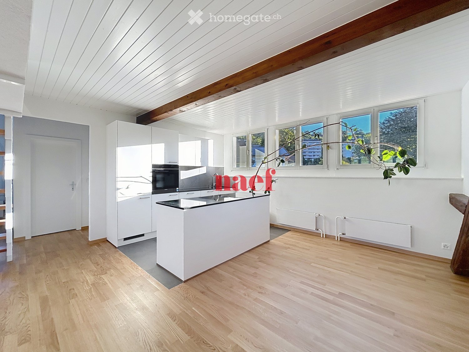 Modern kitchen with white cabinets, a central island, and wooden flooring. Features include an oven, sink, and large windows with a view of greenery.