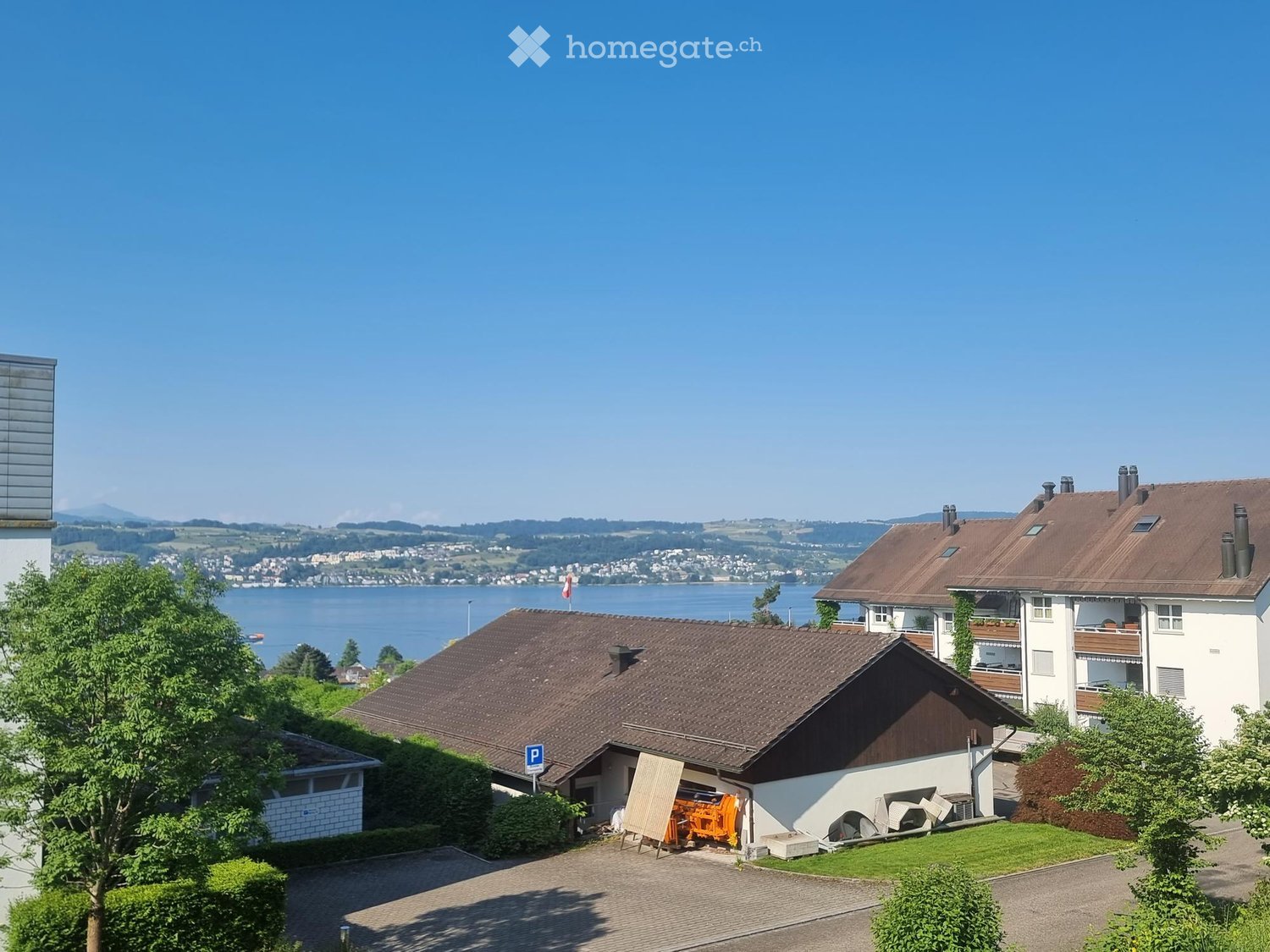 A two-story house with a brown tiled roof, white walls, and a garage. The house is surrounded by greenery and has a scenic view of a lake and distant mountains.