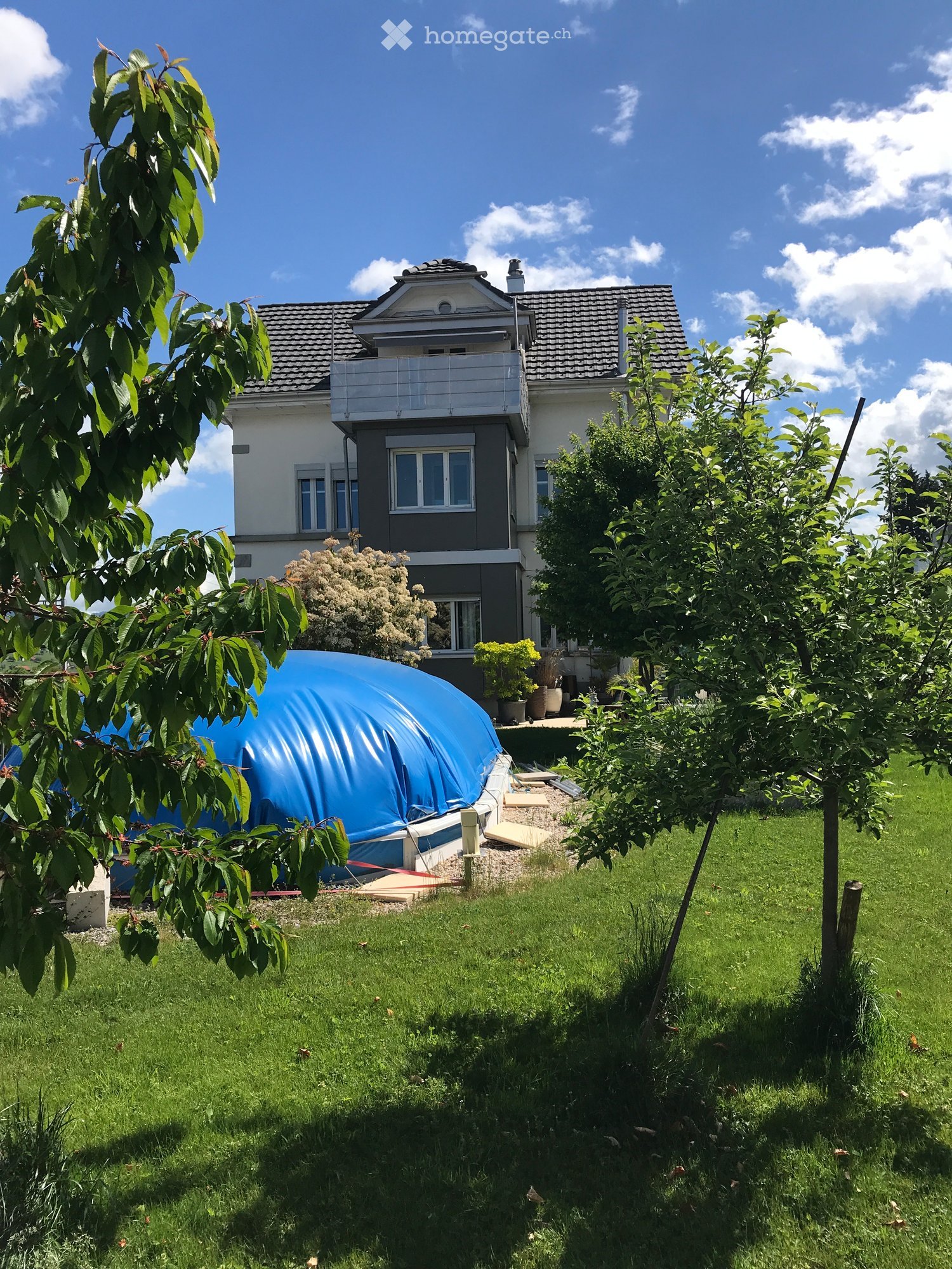 A two-story house with a balcony, surrounded by a well-maintained lawn, trees, and a blue inflatable pool covered with a tarp.