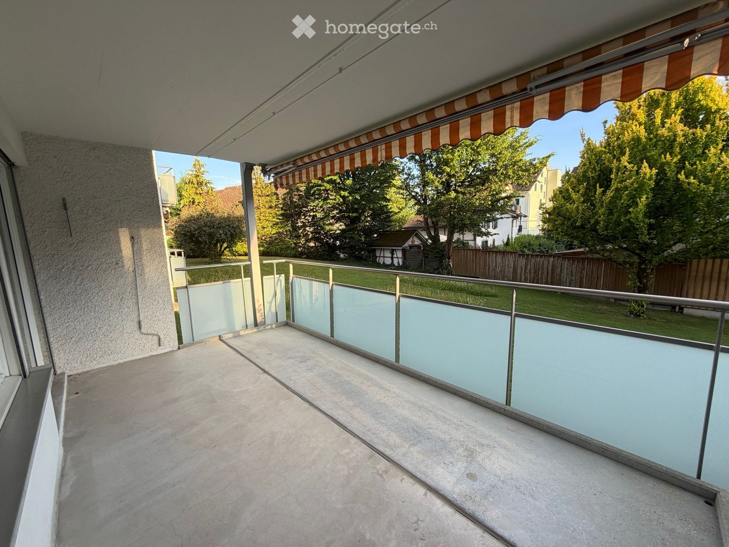 Balcony with concrete floor, glass railing, and striped awning, overlooking a green garden and trees.