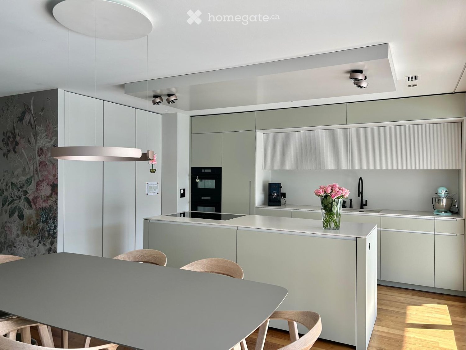 Modern kitchen with light grey cabinets, a central island, and a dining area. Features include a coffee maker, mixer, vase with flowers, and a sleek faucet.