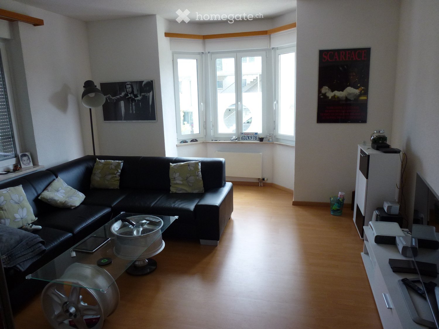 Modern living room with a black leather sofa, glass coffee table, floor lamp, TV stand, and artwork on the walls.