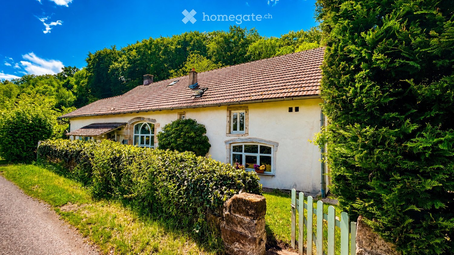 A charming single-story house with a tiled roof, white exterior, and multiple windows, surrounded by lush greenery and a gravel pathway.