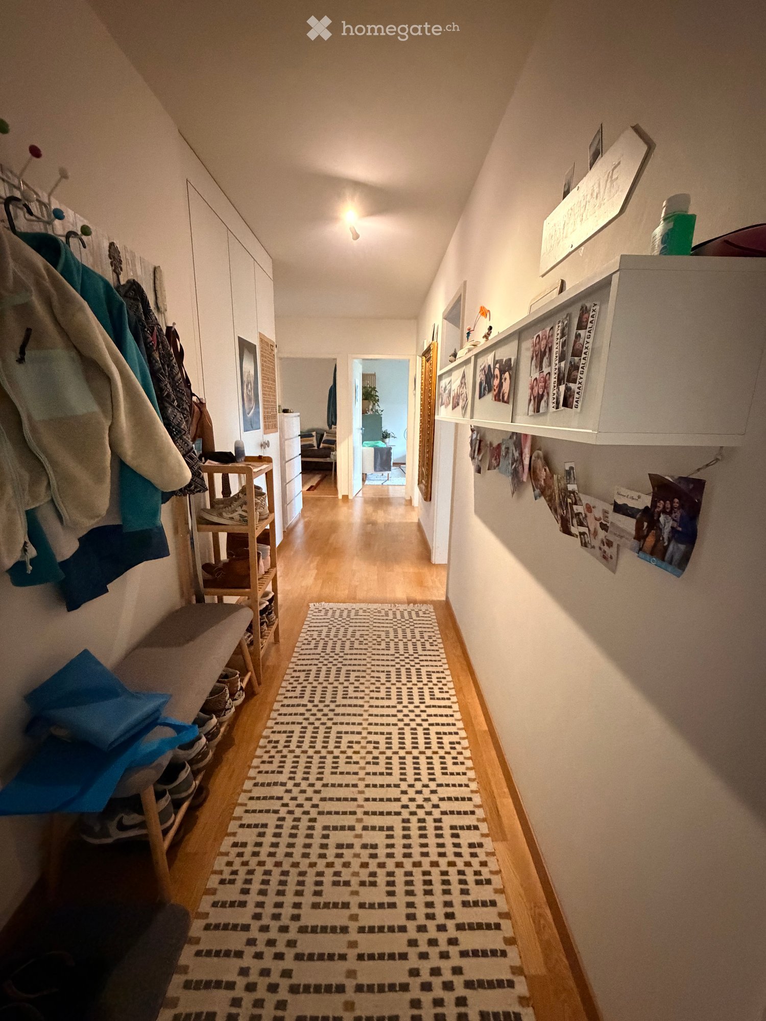 Hallway with wooden flooring, patterned rug, coat rack, shelves, and framed photos on the wall.