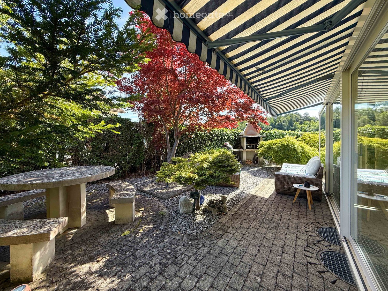 Outdoor patio with stone table, benches, wicker sofa, small table, and a brick fireplace surrounded by greenery and trees.