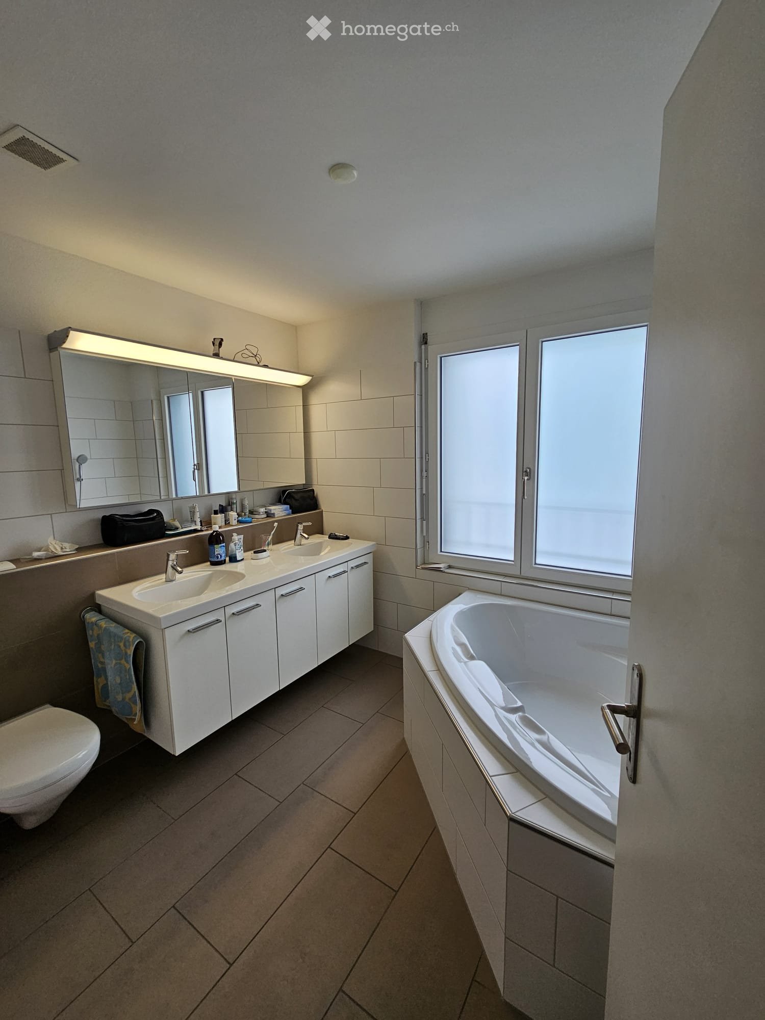 Modern bathroom with a double sink vanity, large mirror, bathtub, and toilet. The space features tiled walls and floor, and has a window providing natural light.