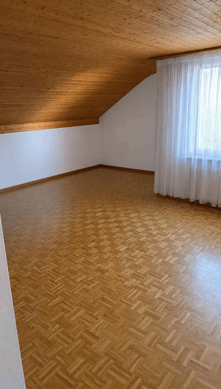 Empty room with wooden herringbone flooring, white walls, and a slanted wooden ceiling. A large window with white curtains allows natural light to enter.
