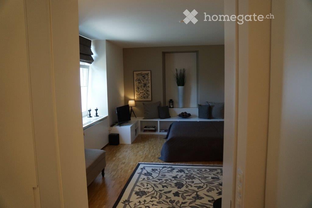 Cozy bedroom with a large bed, nightstand, lamp, television, and a patterned rug on the floor. A window with blinds and a small seating area near the window are also visible.