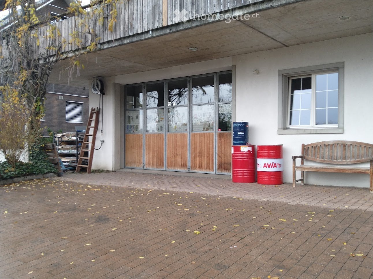 Outdoor area with brick patio, wooden bench, red and blue barrels, and a wooden ladder leaning against the building.