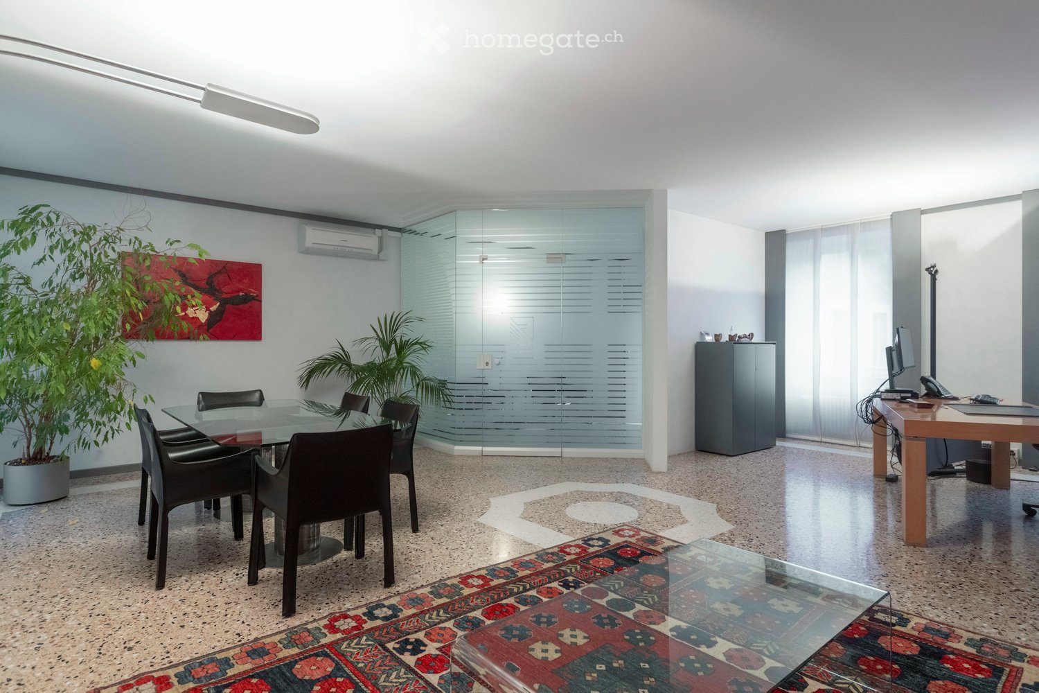 Modern office space with a glass dining table and black chairs, a wooden desk with a computer monitor, a cabinet, a potted plant, and a painting on the wall.
